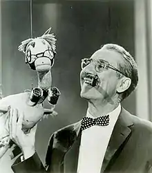 Photo en noir et blanc d'un homme moustachu à lunettes, souriant, un cigare entre les dents et tenant dans sa main droite un canard en peluche lui ressemblant