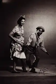 Photographie en noir et blanc, une femme est debout à gauche et à droite un homme est légèrement accroupi. La femme est vêtue d'une robe et l'homme d'un pantalon et d'une chemise.