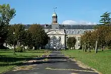 Vue de face d'un bâtiment à deux étages, dont un sous le comble brisé. Le corps central est surmonté d'une lanterne. Au premier plan, pelouses et arbres de la cour.