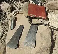Posés sur une pierre plate, au premier plan, deux lames ou grattoirs en métal, au deuxième plan, un autre outil rattaché à une corde et une besace en cuir.