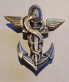 Insigne militaire porté sur la poitrine représentant une ancre de marine sur laquelle s'enroule le serpent d'Esculape, l'anneau supérieur de l'ancre est remplacé par le miroir de la sagesse