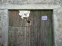  Une porte de grange en bois sombre située dans le hameau de Recoulette, fermée par des targettes en métal et entourée par des piliers et un linteau en béton. En haut à gauche, un morceau de tissu blanc plié dépasse de la porte. En haut à droite, un panonceau bleu avec un texte en blanc : "Département de la Lozère. Services vétérinaires. Étable indemne de tuberculose".