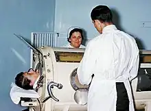 À la gauche, la tête d'une personne émerge d'un appareil. Deux personnes se tiennent debout de part et d'autre de l'appareil