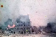 photographie en couleurs montrant des immeubles en ruines dont un encore en flammes