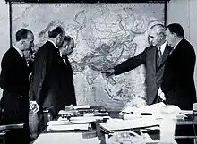 Photo en noir et blanc montrant cinq hommes en costume regardant une carte murale de l'Asie. L'un des hommes pointe la région du Bangladesh à l'Est de l'Inde. Au premier plan, un bureau est recouvert de documents.